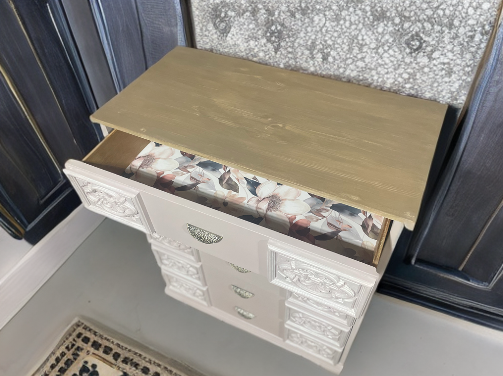 An upcycled or refinished dresser in white with ornate drawer fronts, a natural wood tone top, and the top drawer open to show a floral patterned liner.